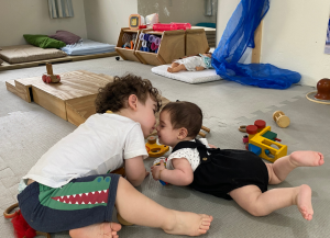 Dois bebes com os rostos próximos, quase tocando o nariz um do outro, em uma demonstração de afeto, uma menina de 8 meses deitada no chão e um menino de 15 meses sentado e inclinado em direção a ela.